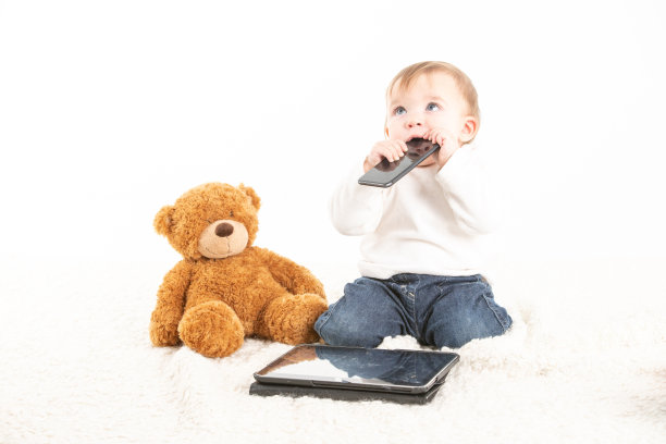 白色背景的工作室照片，一个婴儿的脸，嘴里有一个手机，旁边是一个泰迪熊和一个平板电脑图片下载