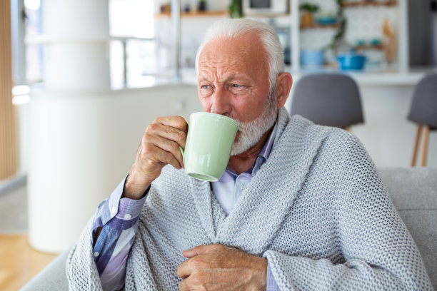 患流感的老人裹着毯子坐在家里的沙发上喝茶。患感冒的病人端着一大杯热茶躺在沙发上。图片下载
