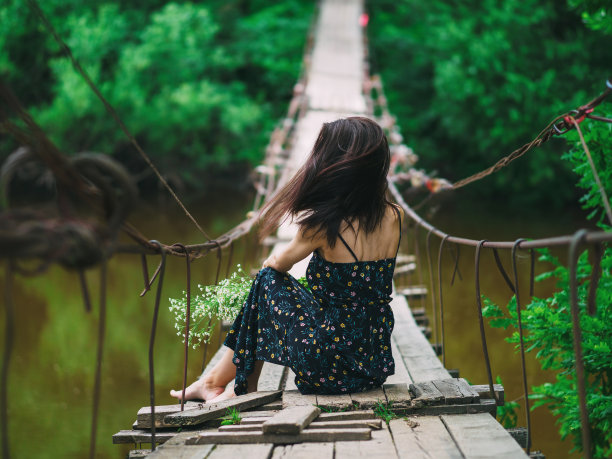 一个女孩穿着夏装，手捧一束雏菊，坐在河上的吊桥上。享受夏日户外。后视图图片下载