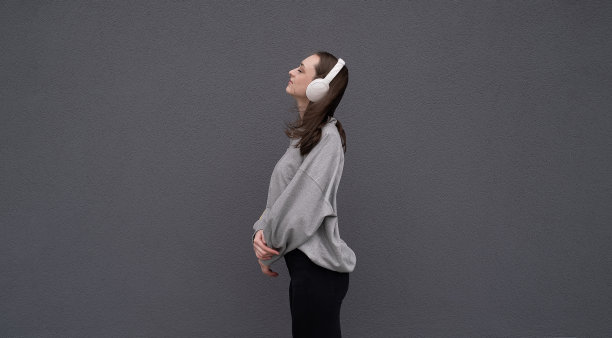 无忧无虑迷人的黑发女孩，穿着灰色毛衣，戴着耳机听音乐，站在灰色的背景上图片下载