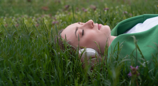 一个十几岁的女孩戴着耳机听音乐，躺在公园的绿草地上，微笑着露出舌头，穿着白色t恤和绿色毛衣，夏天的心情是一种生活方式图片下载