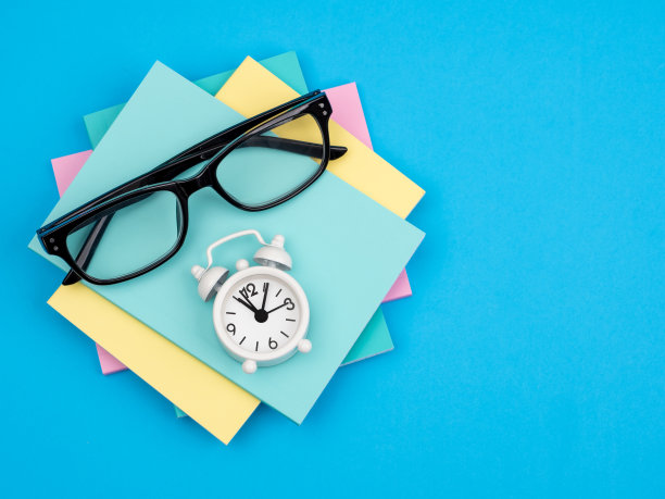一叠用来写字的纸，一副眼镜和一个蓝色背景的闹钟，作为计划和时间管理的概念图片下载