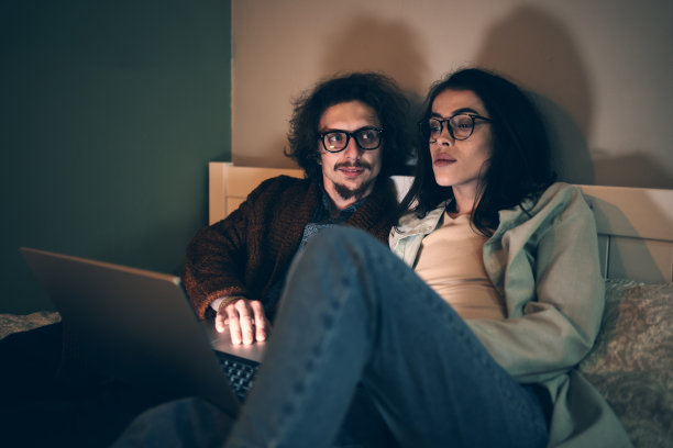 年轻夫妇在家里的沙发上使用笔记本电脑。丈夫和妻子一起网购，看新闻或社交媒体图片下载