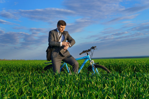 一个商人骑着自行车，拿着公文包，在绿色的草地上看手表，穿着西装，春天的美丽自然，自由职业的商业理念图片下载