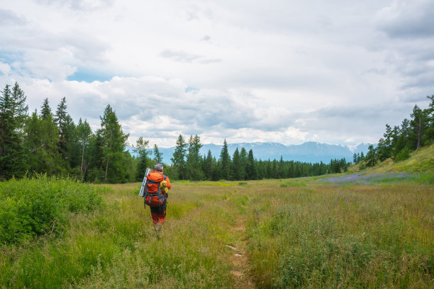 穿着红色衣服背着大背包的旅行者在低云中向高山行进。背包客在多云的天空下穿过森林来到大山。引人注目的景观与游客在山上。图片下载