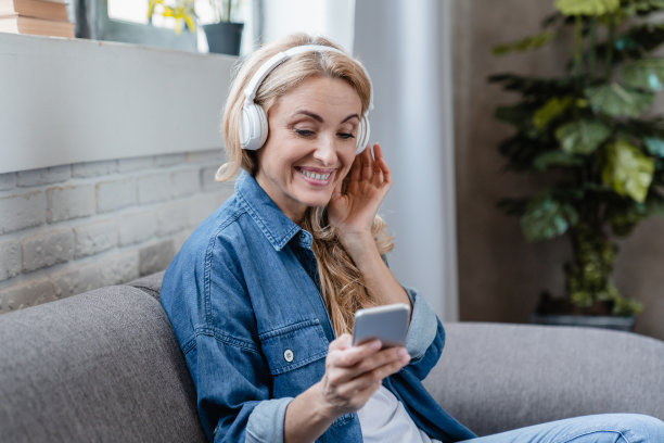 成熟的中年白人女性自由职业者，戴着耳机听音乐广播播客电子书，在家上网用手机选择音乐图片下载