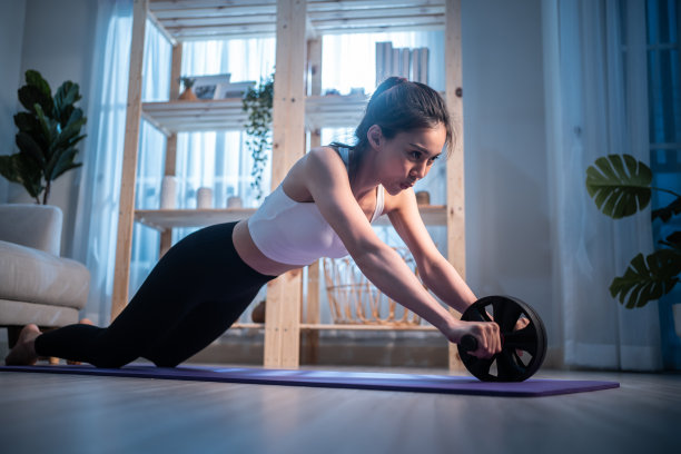 亚洲年轻美丽活跃的运动妇女在家里做有氧运动。迷人的女孩穿着运动服，用abs滚轮在家里的客厅里进行休闲健身。图片下载