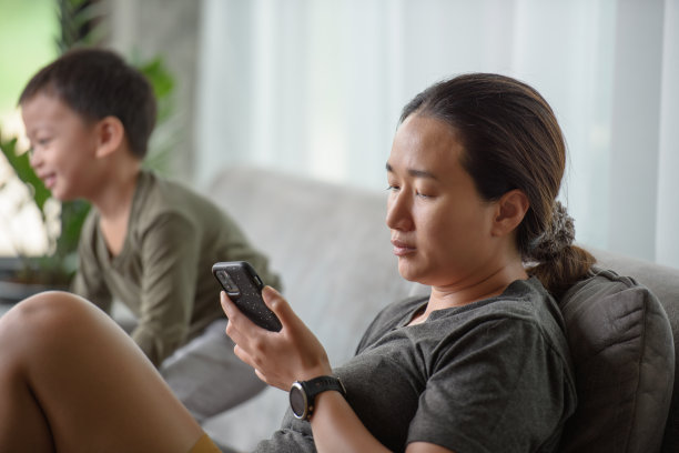 亚洲单身妈妈头痛，感到累，烦吵闹的孩子在家里玩，妈妈通过智能手机在线工作。不要在意孩子和孩子玩电话。图片下载