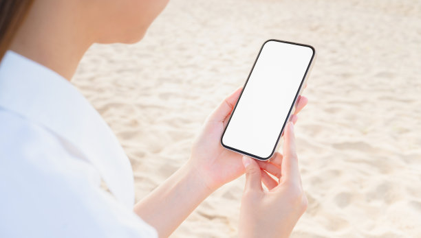 手持智能手机在沙滩上，手机模型的空白屏幕，拿你的屏幕投放广告。暑假概念。图片下载