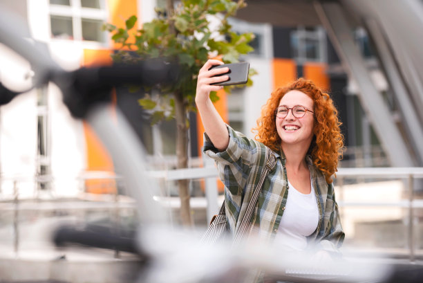 美丽微笑的红发女学生在大学大楼前用智能手机自拍图片下载
