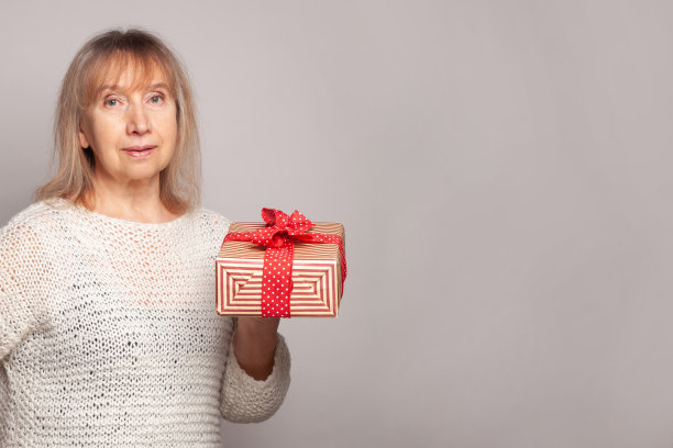 迷人的成熟女人在白色毛衣持有条纹红白礼品盒在灰色旗帜的背景图片下载