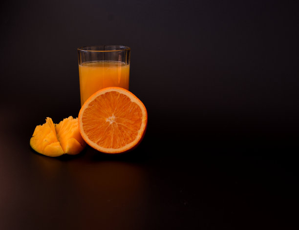 一杯黑色背景的鲜榨果汁，旁边是几片熟芒果和半个橙子。图片下载
