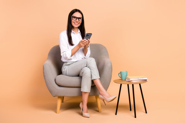 全身照片可爱的微笑的女人穿着白衬衫坐在椅子上打字现代设备孤立的米色背景图片下载