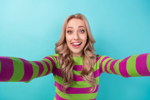 女孩的肖像华丽可爱的女孩波浪发型，穿着时尚的衣服，使自拍舌头舔嘴唇孤立的蓝色背景图片下载