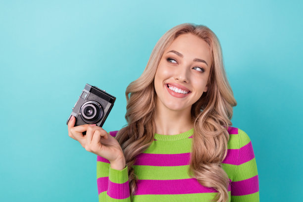 照片中狡猾狡猾的女人卷发，穿着条纹运动衫，拿着相机，在蓝绿色的背景上看起来空无一物图片下载