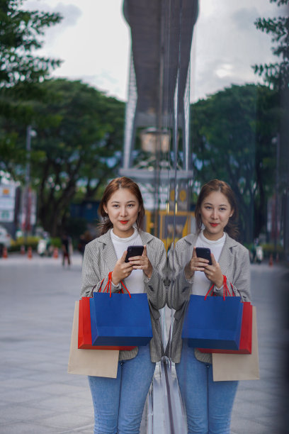 在商场购物时使用你的智能手机。一个漂亮的女孩在购物中心前做很多购物，享受节日购物后自己的肖像。图片下载