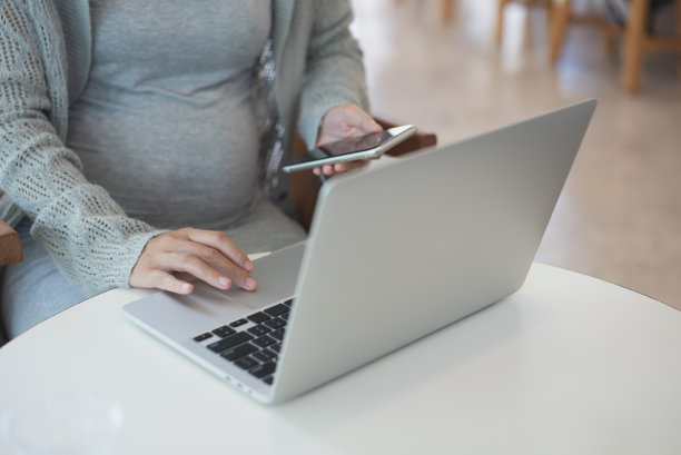 怀孕的女商人坐在桌边玩手机，在咖啡馆用笔记本电脑工作。孕妇通过手机app网购，近距离图片下载