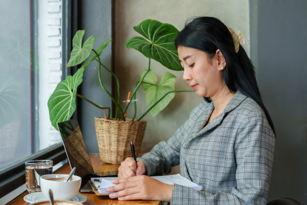 亚洲女商人交易投资组合坐在桌子靠窗的手拿着笔和检查文件躺在桌子上在笔记本电脑前的女人在办公室外工作图片下载