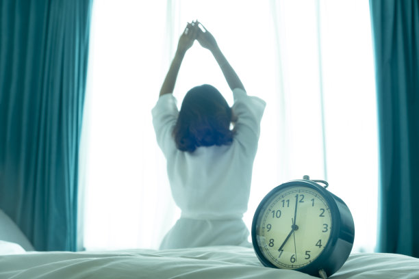 新的一天的早晨，闹钟叫醒了坐在房间里的女人。一位妇女在窗口伸展肌肉。健康和护理概念图片下载