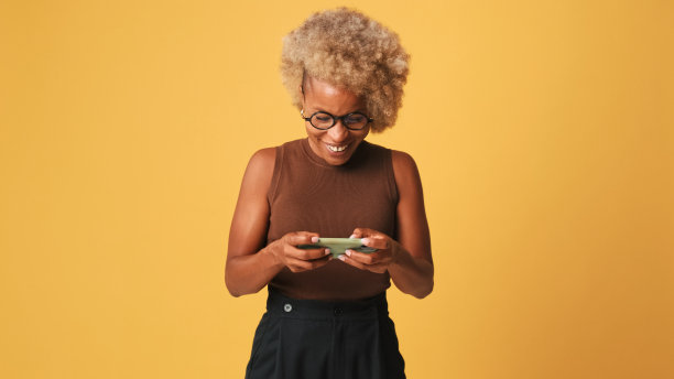 快乐的女孩戴着眼镜，穿着棕色上衣，正在使用手机，在橙色的背景上孤立地发短信图片下载