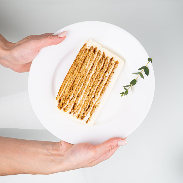裁剪的观点的女人持有一块蜂蜜蛋糕在一个白色的盘子。把蛋糕切成看得见的层次和白色奶油图片下载