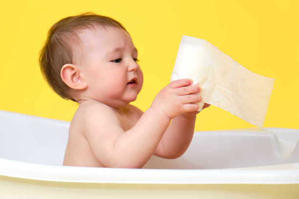 好奇的婴儿蹒跚学步的男孩与卫生纸在一个白色的浴缸，工作室的黄色背景。一岁快乐的孩子图片下载