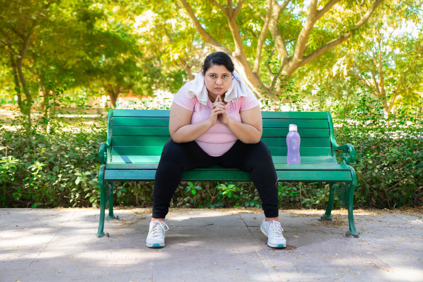 一名超重的印度妇女穿着运动服，脖子上围着毛巾，悠闲地坐在夏日公园户外的长凳上。健康的生活方式。全裸镜头。图片下载