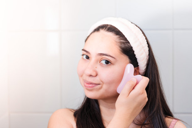 微笑的年轻女子在镜子里用瓜沙石英石刮刀按摩她的脸颊。图片下载