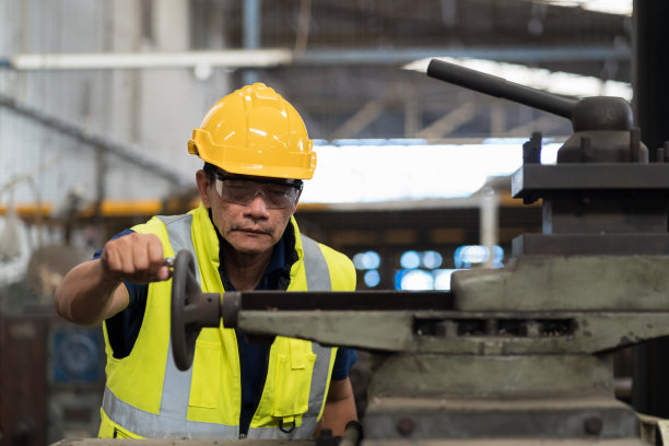 在工业工厂检查车床零件质量的资深亚洲男工程师，穿着安全制服，头戴安全帽。成熟的男性技术工人维护机器零件图片下载