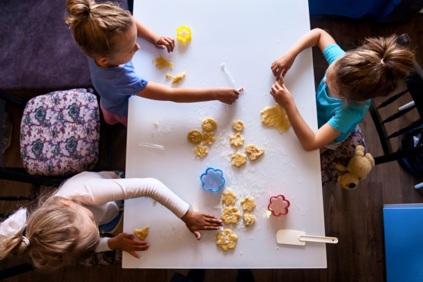俯视图，三个小女孩坐在厨房的桌子上，在家里用面团和面粉做饼干。孩子们在一起给家庭带来惊喜。享受烹饪过程概念。复制广告文本空间图片下载