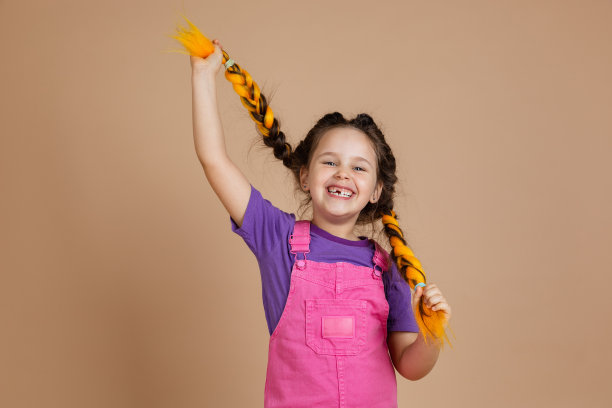 快乐兴奋的小女孩，扎着黄色的kanekalon辫子，露出灿烂的笑容，看着镜头，穿着粉红色的连体衣和紫色的t恤，米色的背景图片下载