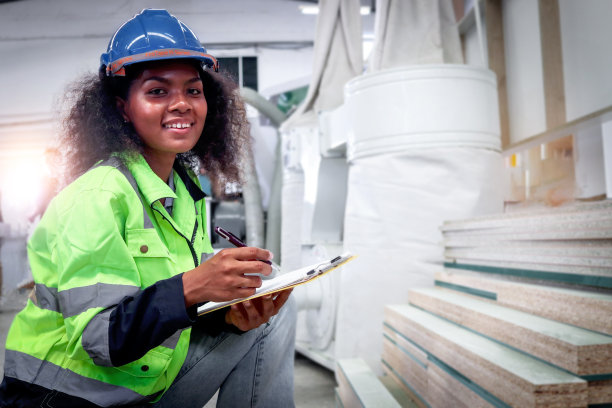 快乐微笑的卷发非洲女工戴着头盔和安全背心，在文件的文件夹上写字，在家具制造厂检查木材原材料，数控木工图片下载