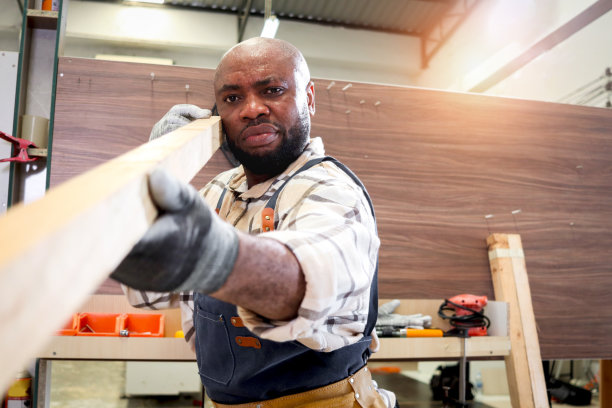 非洲中年木匠系着围裙，测量瞄准木板的人像，木工木工车间制作木制家具的工人人像，木工车间做工的工匠人像。图片下载