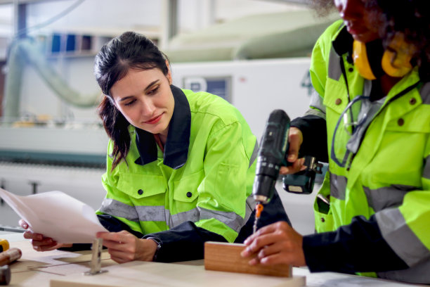 穿着安全背心的工业美女工人拿着图纸，和非洲卷发手工打钻的女同事商量，一起在数控木工家具厂工作图片下载