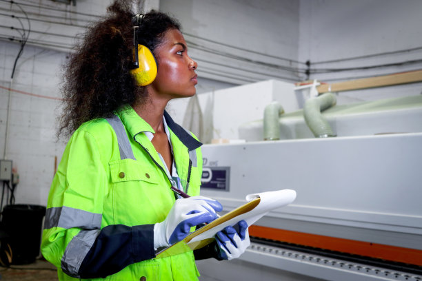 卷发的非洲工业女工穿着安全背心，在文件夹上写文件，女工程师在数控木工制造家具厂检查和维修机械。图片下载