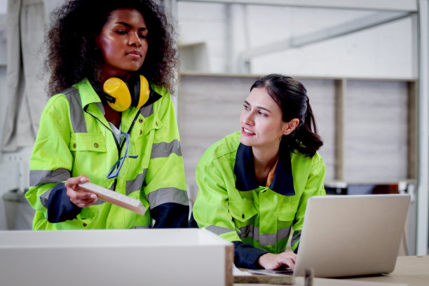 在数控木工家具厂，穿着安全背心的美丽工业女机械工程师一边用笔记本电脑操作机械，一边与非洲卷发女同事讨论。图片下载