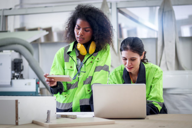 在数控木工家具厂，穿着安全背心的美丽工业女机械工程师一边用笔记本电脑操作机械，一边与非洲卷发女同事讨论。图片下载