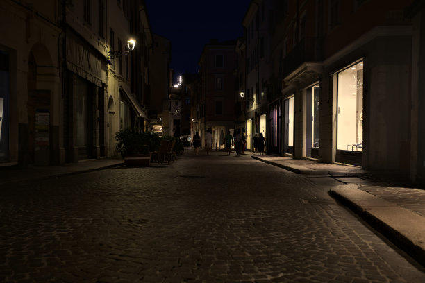 意大利小镇夜晚铺满鹅卵石的街道上有商店橱窗和餐馆的桌子图片下载