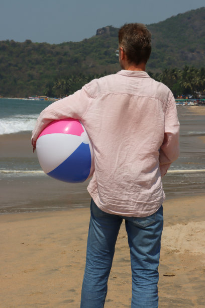 近距离照片:一名无法辨认的白人男子身穿牛仔裤和长袖衬衫，站在泻湖水边的沙滩上，腋下夹着沙滩球和色彩鲜艳的沙滩玩具，后景是长满棕榈树的林地图片下载