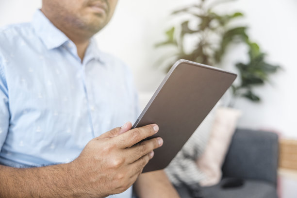 穿着蓝色衬衫的亚洲人坐在客厅的沙发上玩平板电脑。假期放松。在线购物。现代通信技术。关于科技，休息，购物，放松。图片下载