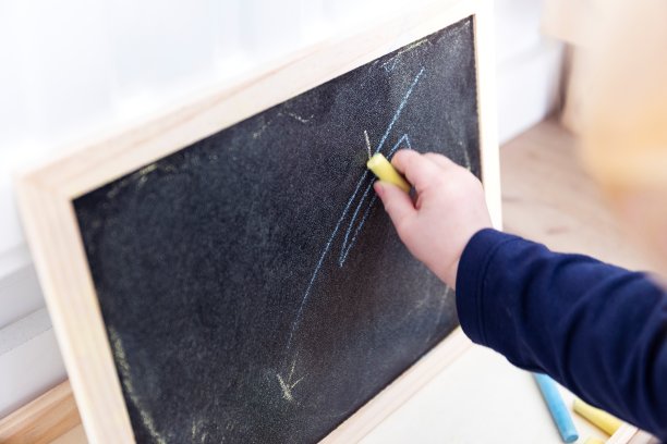 一个蹒跚学步的孩子在黑板上画画、乱涂乱画的特写。这个孩子正在黑板上写字，手里拿着一支黄色的粉笔。图片下载