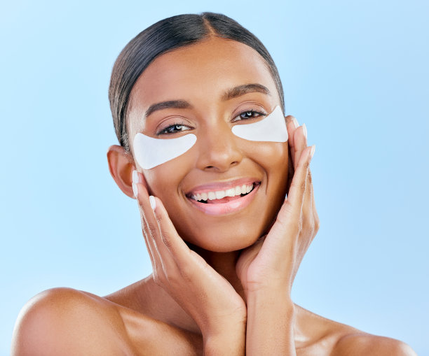 发光，护肤和女性肖像与眼罩，黑眼圈和治疗。微笑，脸和女孩或模特与皮肤科面部胶原蛋白面膜隔离在蓝色背景的工作室图片下载