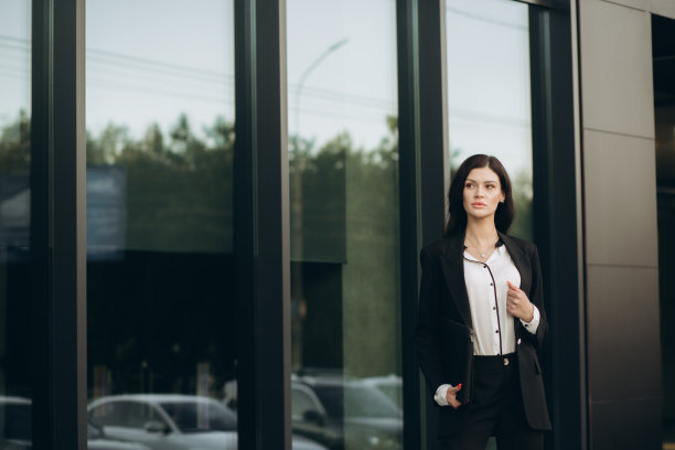 一位身穿黑色西装的年轻女子喝着咖啡，拿着笔记本电脑在街上摆姿势。商业肖像图片下载