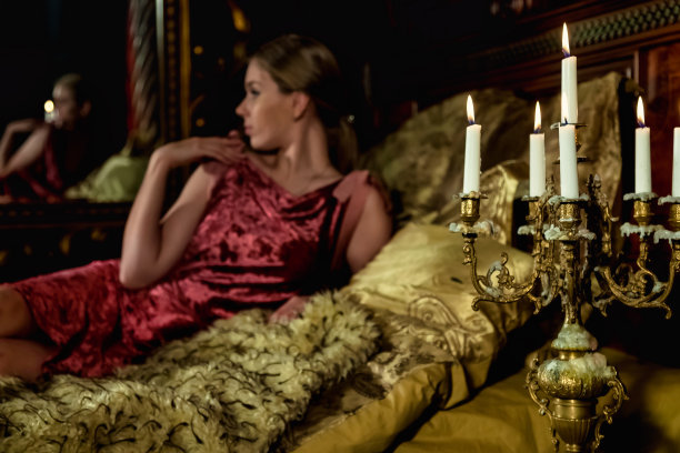 可爱的女人在时尚的形象摆姿势躺在床上在中世纪的卧室，选择性的焦点和烛台。沉思的年轻女士在室内古色古香的金色房间里。剧场表演的概念。复制广告文本空间图片下载