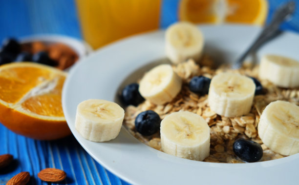 燕麦片配上新鲜的浆果，香蕉放在白色盘子里，配上橙汁，蓝色的木制背景。图片下载