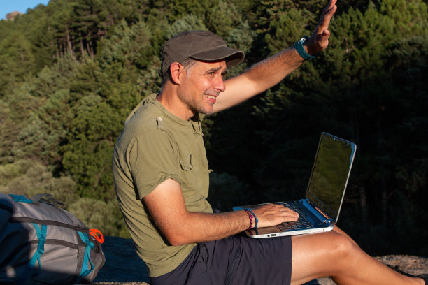 戴着帽子的男人坐在灌木丛中的一块石头上，他带了一台笔记本电脑，可以在大自然中工作图片下载