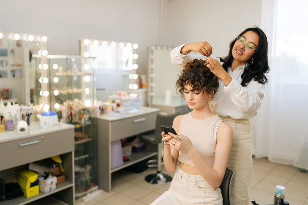 优雅的年轻女性客户坐在美容院的椅子上使用智能手机，而美发师则在美容院提供服务和护理。图片下载