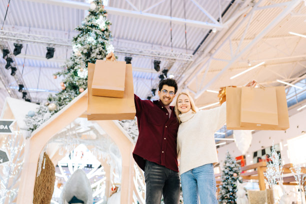 在购物中心的大厅里，一对幸福的年轻夫妇拿着装满礼物的袋子，站在一起拥抱着镜头，从低角度看他们一起享受购物图片下载