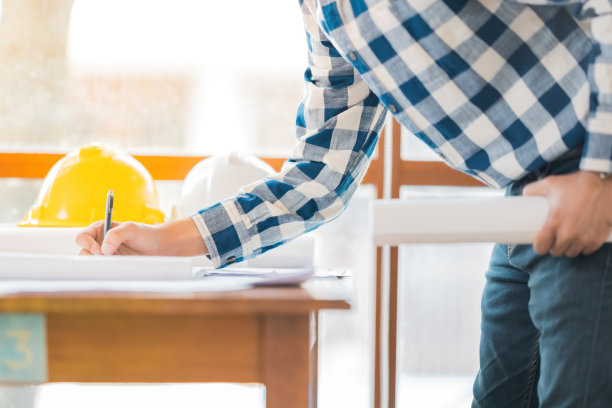 一名男性工程师用笔在蓝图中设计结构的手持照片。他旁边的桌子上放着一顶黄白相间的头盔。在办公室工作场所穿着蓝色格子制服图片下载