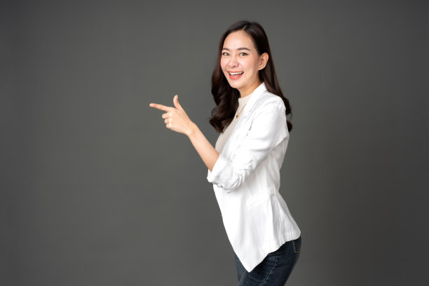 长发亚洲女性高管侧身，手指指向目的地。穿着白色西装和牛仔裤，兴奋的脸上，灿烂的笑容。在灰色背景的摄影棚里拍一张照片图片下载